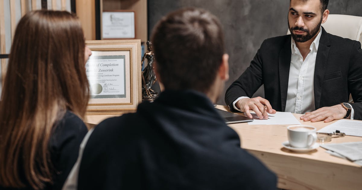A lawyer reviews documents with two clients across a desk in a warm professional office with a Lady Justice statue visible on the bookshelf